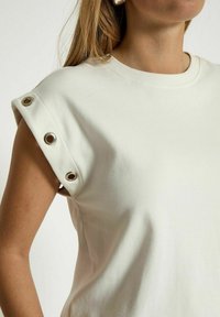Camicia corta beige chiaro a maniche corte con collo tondo; presenta dettagli a occhio sulla spalla. Tessuto liscio, vestibilità casual, design minimalista.