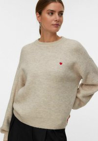 Donna che indossa un maglione di maglia beige con un piccolo cuore rosso sul petto, abbinato a pantaloni neri, che guarda di lato con le mani sui fianchi.