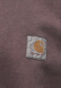 Tessut tricotat de culoare violet închis, cu un etichetă rectangulară din țesătură gri, pe care este imprimat un logo galben și textul "carhartt" brodat în culoarea vișinie.