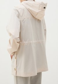 Chaqueta ligera de color beige con capucha, puños elásticos, detalles de botones en los laterales y un corte relajado. Textura suave, diseño minimalista.