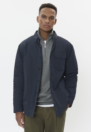 Navy button-up jacket over a gray zip-neck shirt, with a chest pocket and contrasting collar. Slim fit, textured fabric, minimal design.