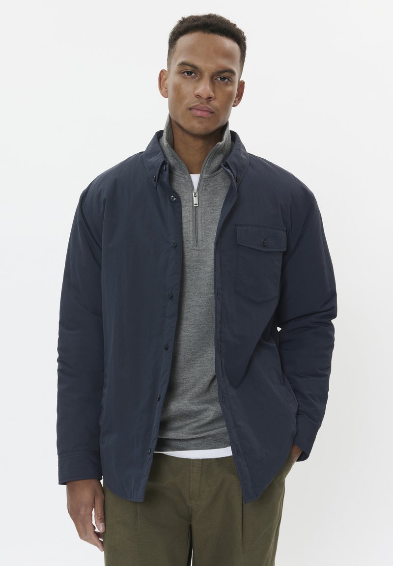 Veste boutonnée bleu marine sur une chemise à col zippé gris, avec une poche poitrine et un col contrastant. Coupe ajustée, tissu texturé, design minimal.