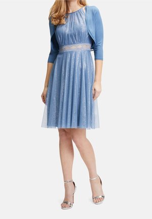 Kvinde iført en glitrende blå knælængde kjole med gennemsigtig taljedetalje og en matchende kort blå cardigan, kombineret med sølvfarvede højhælede sandaler.