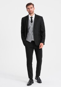 Mustat puku harmaalla liivillä, valkoisella paidalla ja mustalla solmiolla. Slim fit -housut ja mustat kengät täydentävät asukokonaisuuden. Sileä kangas, jämäkkä muotoilu.