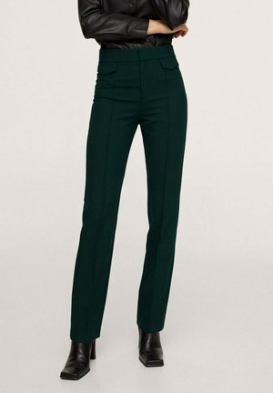 Vrouw die donkergroene op maat gemaakte broek draagt met voorvakken met klep, zwarte laarzen met hak en een zwart leren jasje met gekruiste armen.