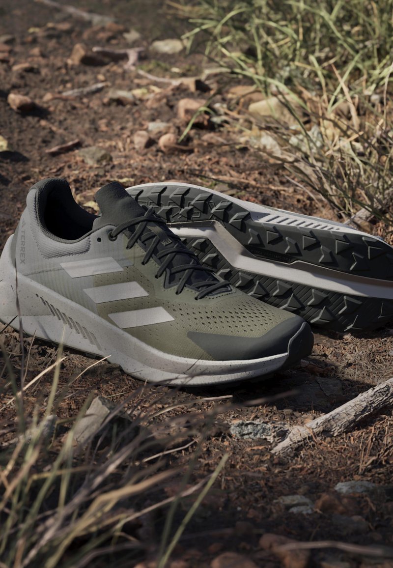 Green athletic shoes with a textured upper, perforations, and black laces. Features a thick sole with a rugged tread pattern and branding details.