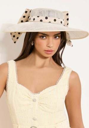 Junge Frau mit einem breitkrempigen beigen Hut mit schwarzem gepunktetem Band und einem blassgelben ärmellosen Kleid mit dekorativen Knöpfen.