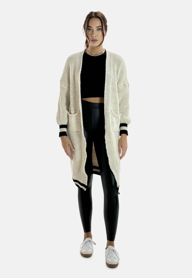 Krämfärgad lång kofta med ribbad textur, svarta accenter och två framfickor, kombinerad med svarta leggings och vita sneakers.