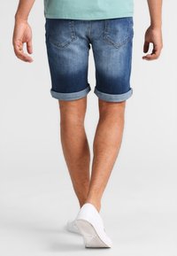 Pantalones cortos de mezclilla en un azul degradado con puños enrollados. Presentan dos bolsillos traseros y un aspecto desgastado, combinados con zapatillas blancas.