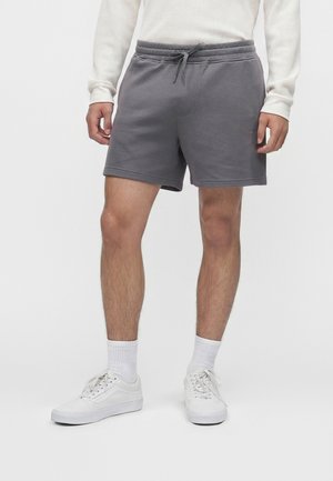 Homme portant un short gris à cordon, un haut blanc à manches longues, des chaussettes blanches et des baskets blanches sur un fond uni.