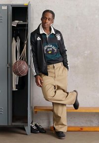 Μαύρο αθλητικό μπουφάν με λευκές λεπτομέρειες, πράσινη μπλούζα με κείμενο, μπεζ παντελόνι, μαύρα παπούτσια. Ντουλάπα στο φόντο με μπάσκετ και ρούχα.