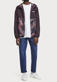 Vindjacka i mörk lila med ett svart och rosa palmbladsmönster. Har en dragkedja fram, justerbar huva och elastiska ärmslut.