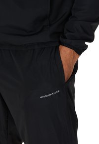 Svart idrottsjacka och byxor med elastiska manschetter, sidofickor och "ENDURANCE"-logga längst ner till vänster på byxorna. Mjuk textur.