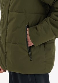 Groene, gevoerde jas met een ritszak aan de linkerkant, gemaakt van gladde stof met een matte afwerking en minimale stikdetails.