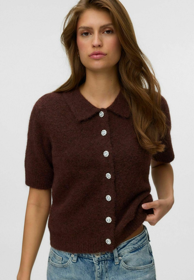 Femme aux longs cheveux bruns portant un cardigan court en maille peluche marron foncé avec six boutons décoratifs et un jean bleu clair.