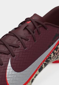 Κοντινό πλάνο ενός μπορντό ποδοσφαιρικού παπουτσιού με σκούρες κορδόνια, ασημί λογότυπο Nike και το κείμενο "Nike United" στη σόλα με κόκκινο και καμουφλάζ σχέδιο.