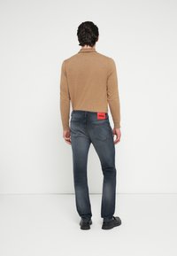 Cămașă maro cu mâneci lungi, blugi din denim albastru închis cu o etichetă portocalie HUGO pe buzunarul din spate, purtate cu pantofi negri.