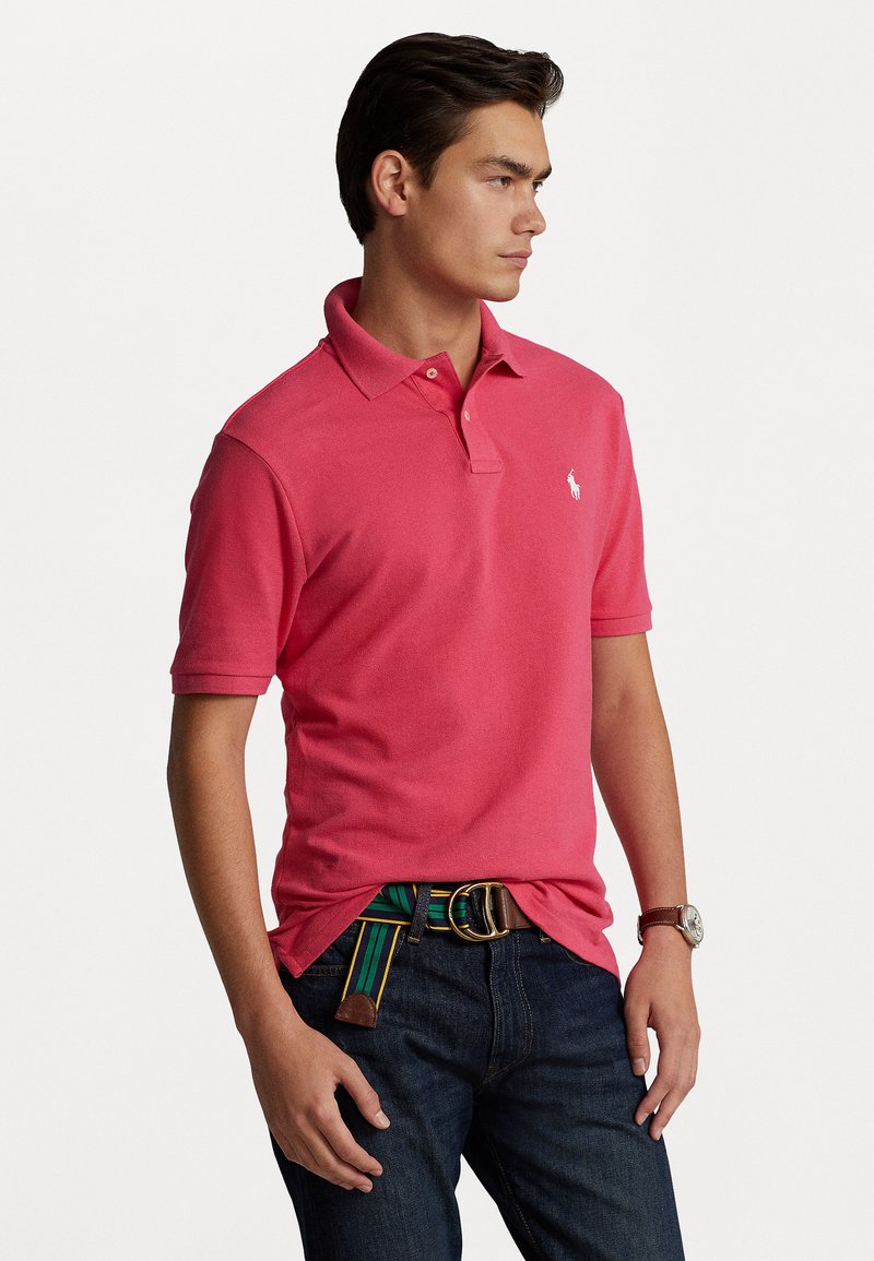 Polo rose en coton, avec un col, des manches courtes et un petit logo blanc. Porté avec un jean foncé et une ceinture multicolore.