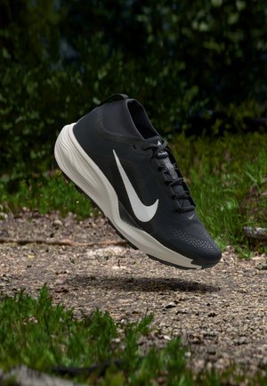 Sort og hvid Nike løbesko, der svæver over en grussti omgivet af grønne planter.