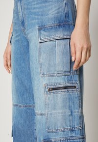 Vaaleansiniset farkkupunokset, joissa on korostetut taskut, vetoketjut ja kontrastivärinen ompelu, rento istuvuus ja pehmeä pinta.