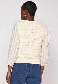 Donna con capelli corti che indossa orecchini a cerchio dorati, un gilet di maglia color crema sopra una blouse a righe e jeans in denim blu, girata di spalle rispetto alla macchina fotografica.