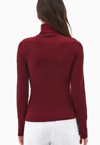 Burgundowa bluzka z długim rękawem, wysokim kołnierzem, prążkowaną teksturą, dopasowanym krojem i zaokrąglonym dołem, noszona z białymi spodniami.