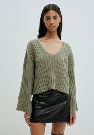 Olivgrüner, gerippter Strickpullover mit V-Ausschnitt und weiten Ärmeln, kombiniert mit einem schwarzen Leder-Minirock, der einen geschichteten Look erzeugt.