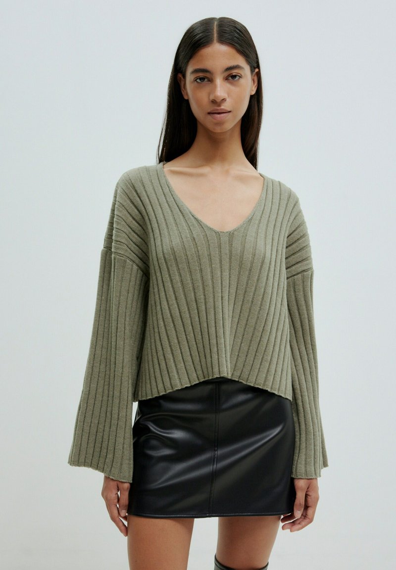 Olivgrüner, gerippter Strickpullover mit V-Ausschnitt und weiten Ärmeln, kombiniert mit einem schwarzen Leder-Minirock, der einen geschichteten Look erzeugt.