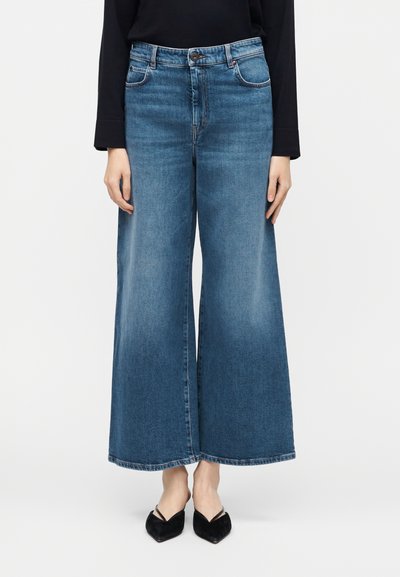 Jean en denim bleu à jambes larges porté avec des ballerines noires à bout pointu et un haut noir à manches longues, visible de la taille aux pieds.