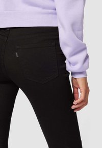 Jeans negros con un diseño ajustado, que presentan un bolsillo trasero característico con un patrón de costura curvo. Acompañados de un sudadera de color púrpura claro.