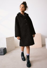 Manteau noir et duveteux avec une capuche, doté d'une fermeture à boutons et de manches larges. Associé à des bottines noires et des chaussettes violet clair.