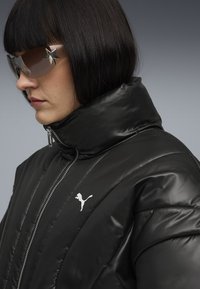 Zwarte pufferjack met hoge kraag, geborduurd textuur, zilveren rits en wit Puma-logo. Model draagt oversized, getinte zonnebril.