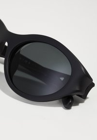 Lunettes de soleil en plastique noir avec des verres ronds, finition mate et un design minimaliste. Elles possèdent des branches courbées et un détail de logo discret sur le verre.