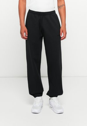 Persoon in zwarte sweatpants met elastische manchetten en trekkoord in de taille, met witte sneakers, staande tegen een simpele witte achtergrond.