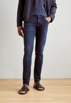 Jeans aderenti in denim blu scuro, con il classico design a cinque tasche. Abbinati a sandali neri e a un top blu navy sotto una giacca.