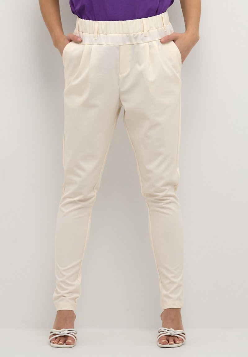 Pantalon fuselé de couleur crème avec une ceinture élastique, doté de poches latérales et d'un tissu lisse et léger. Porté avec des sandales à brides.