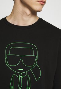 T-shirt nera con una grafica verde neon di un personaggio con occhiali da sole e cravatta, realizzata in morbido cotone con una finitura testurizzata.