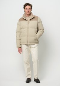 Beżowa kurtka puchowa z akcentem z sztruksu na ramionach, z zamkiem błyskawicznym i bocznymi kieszeniami, noszona z jasnymi spodniami i ciemnymi butami.