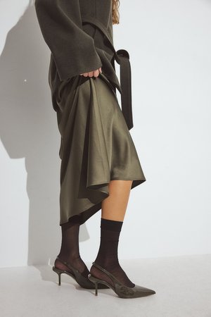 Olive green satin skirt draped asymmetrically; paired with sheer black socks and pointed-toe green heels with a glossy finish.