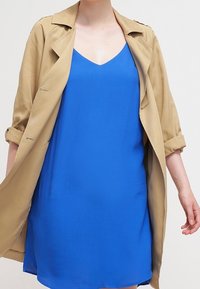 Gabardina beige sobre un vestido sin mangas de un azul profundo. El vestido tiene un escote en V y una textura suave y fluida.