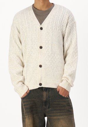 Cárdigan de punto trenzado color crema con escote en V, que presenta patrones texturizados y botones oscuros, combinado con unos vaqueros oscuros de corte holgado.