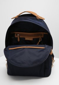 Marin nylon ryggsäck med bruna läderdetaljer, rundade klaffar och flera fack. Har en dragkedjeficka och etikett inuti.