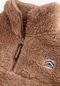 Giacca in pile di un marrone chiaro con una superficie texutizzata e morbida. Presenta una cerniera, un'etichetta con logo e un colletto a costine.