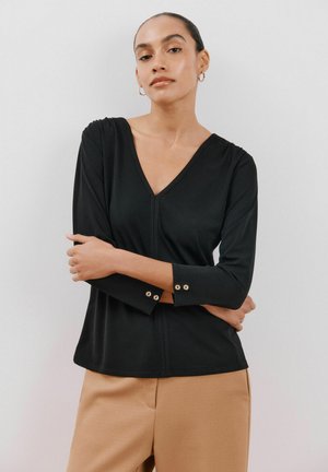 Černý top s výstřihem do V a rukávy třičtvrteční délky, doplněný o knoflíkové detaily. Vyrobený z hladkého materiálu s volným střihem, v kombinaci s světle hnědými kalhotami.
