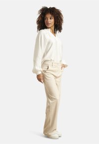 White long-sleeve blouse with embroidered detailing at cuffs, paired with cream-colored trousers and white sneakers, shown in a side profile.