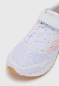 Chaussure de sport blanche avec des accents roses, tige en maille, bout arrondi, semelle plate et lacets visibles. Elle dispose d'une sangle en velcro pour un soutien supplémentaire.