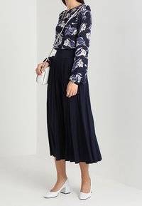 Blusa floreale blu navy con maniche lunghe e scollatura rotonda, abbinata a una gonna midi plissettata blu navy e scarpe con tacco a blocco bianche. Piccola borsa a tracolla.