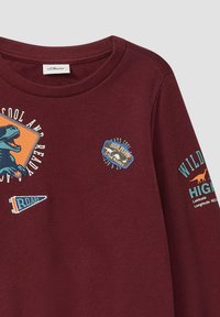 Felpa bordeaux con grafiche di dinosauri, dettagli stampati, colletto a costine e vari etichette colorate sul corpo e sulle maniche.