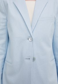 Sacou bleu deschis realizat dintr-un material texturat, având două butoane, revere decupate și un singur buzunar la piept cu detalii subtile de cusătură.