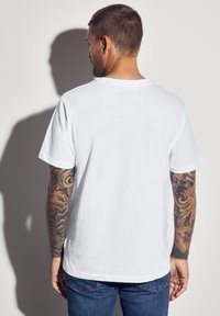 Bílá bavlněná trička s kulatým výstřihem, krátkými rukávy a volným střihem. Výrazné tetování na pažích.